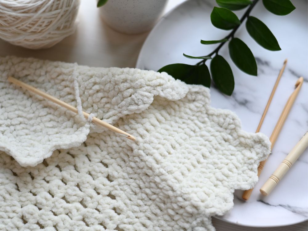 Projeto de crochê inacabado sobre uma mesa com um novelo de lã, um prato de mármore, agulhas de crochê e um ramo de planta.